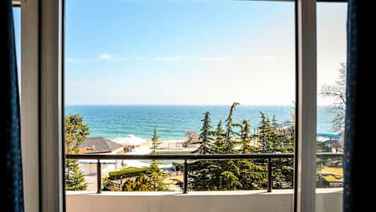 Utsikten gjennom et vindu viser havet, furutrær, hus og en balkong. (AI-generert tekst)