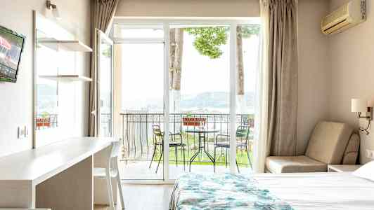 Et hotellrom med en seng, et bord og en stol, samt en balkong med utemøbler og utsikt mot naturen. (AI-generert tekst)