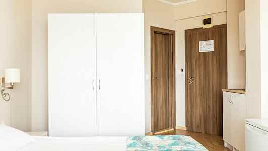 Et hotellromsmiljø med en hvit garderobe, tredører og en del av en seng i forgrunnen. (AI-generert tekst)