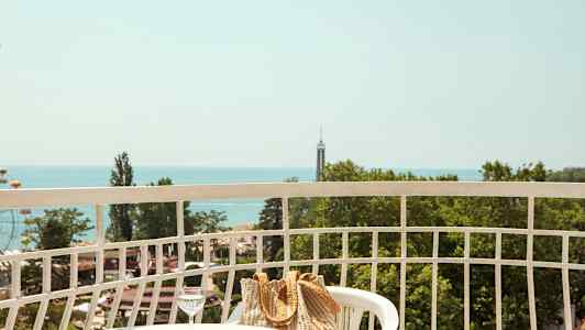 Utsikt fra en balkong med et bord, stoler, en veske og et glass vann, samt grønne trær og havet i bakgrunnen. (AI-generert tekst)