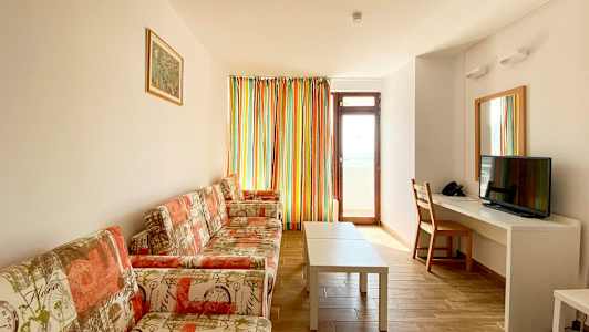 Bilde av en stue med sofa, sofabord, en TV og et skrivebord. En dør og et vindu med fargerike gardiner er synlige. (AI-generert tekst)