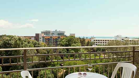 Utsikt fra en balkong med bord og stol, med grøntområder, bygninger og havet i bakgrunnen. (AI-generert tekst)