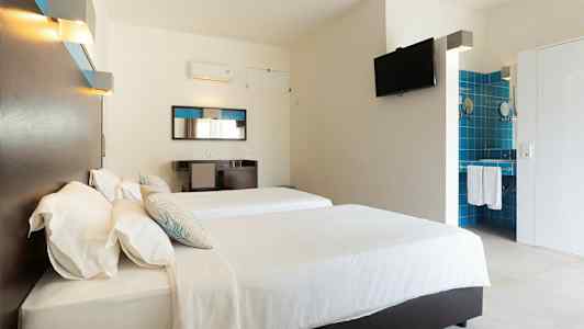 Et hotellrom med to senger, hvitt sengetøy og en veggmontert TV. Speil og skrivebord er plassert på den ene veggen, samt et åpent bad med blå fliser. (AI-generert tekst)