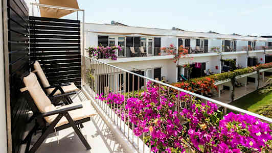 Balkong med utendørsstoler og blomstrende planter foran en bygning med flere leiligheter. (AI-generert tekst)