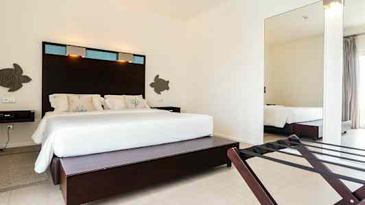 Et hotellrom med en stor seng med hvitt sengetøy og dekorative puter, samt mørkt panel bak sengen. (AI-generert tekst)