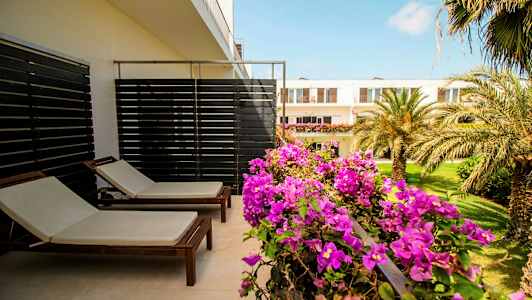 Terrasse med to solsenger og blomstrende planter, med utsikt mot en hage og bygninger. (AI-generert tekst)