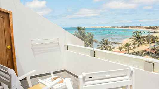 Bildet viser en uteplass med hvite stoler og bord samt utsikt over en strand med palmer, hav og små båter. (AI-generert tekst)