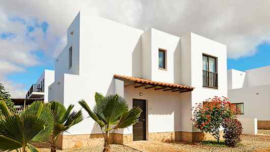 En hvit bygning med en steinsokkel, omgitt av planter og en treinngang. (AI-generert tekst)