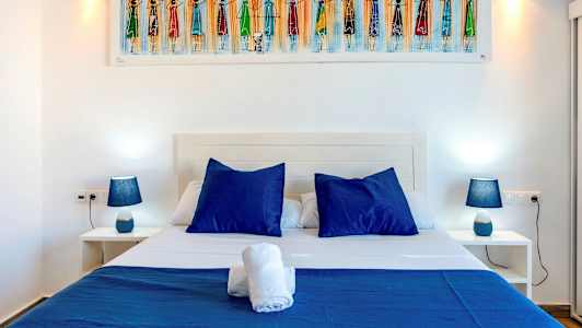 Bildet viser en seng med blå puter og sengetøy, flankert av to nattbord med blå lamper. (AI-generert tekst)