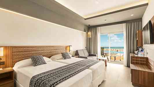 Et hotellrom med to senger og en sittegruppe ved siden av en glassdør som fører til en balkong med utsikt. (AI-generert tekst)