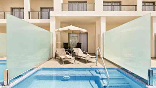 Et bassengområde med to solsenger og en parasoll foran en hvit bygning med balkonger. (AI-generert tekst)