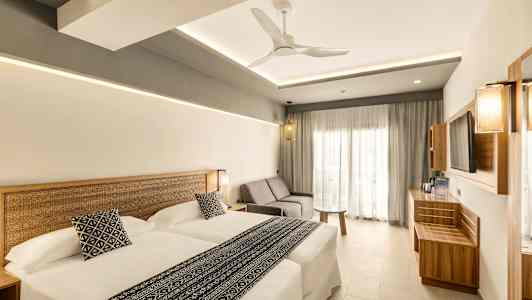 Et moderne hotellrom med to senger, en sofa og et bord. Stilen er lys og minimalistisk. (AI-generert tekst)