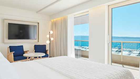 Interiør av et hotellrom med en stor seng, to stoler, et bord og en TV. Utsikt over sjøen gjennom en balkongdør. (AI-generert tekst)