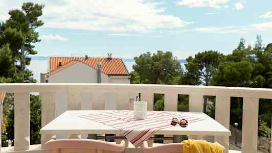 En balkong med bord dekket av stripete duk, solbriller og gul sjal, med utsikt mot havet og hus. (AI-generert tekst)