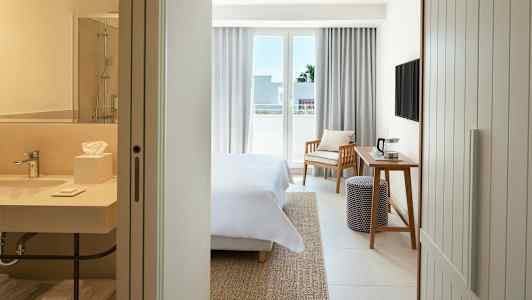 Et hotellrom med en seng, et skrivebord, en stol og en balkong. I forgrunnen ses et baderom. (AI-generert tekst)