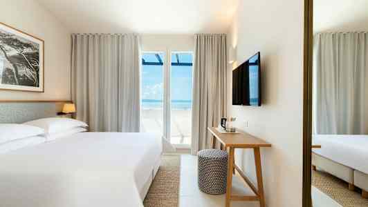 Et hotellrom med en stor seng, et skrivebord og en TV på veggen. Store vinduer med utsikt mot havet. (AI-generert tekst)