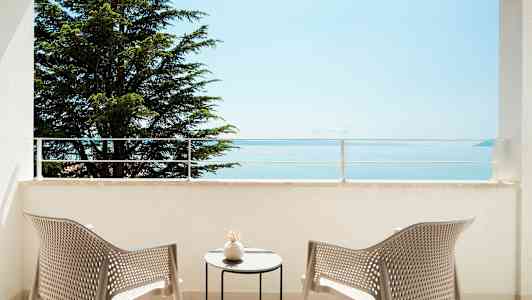 Utsikt fra en balkong med to stoler og et bord, mot sjøen og et tre. (AI-generert tekst)