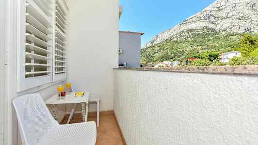 En balkong med to hvite stoler, et bord med to glass juice og frukt samt en utsikt over fjell og omkringliggende bygninger. (AI-generert tekst)