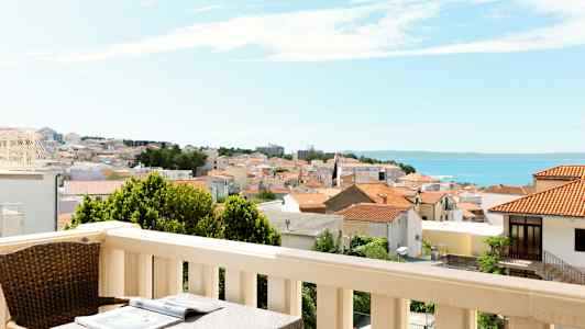 Balkong med bord og stol, utsikt over en by med røde tak og havet i bakgrunnen. (AI-generert tekst)
