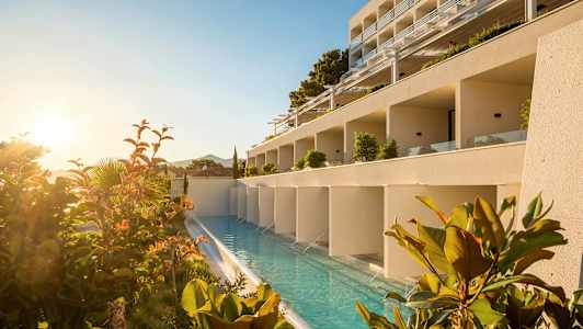 En hotellbygning med et langsgående basseng foran, omgitt av planter og soloppgang. (AI-generert tekst)