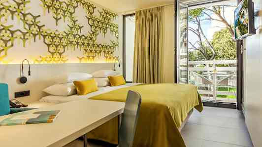 Fotografi av et hotellrom med et gult sengeteppe og dekorativ veggdekorasjon samt utsikt mot et treområde og balkong. (AI-generert tekst)