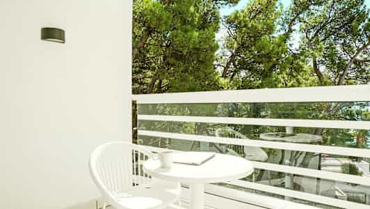 En balkong med et hvitt rundt bord og en hvit stol. Utsikt over grønne trær. (AI-generert tekst)