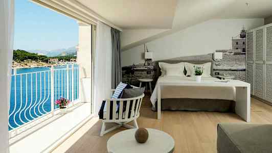 Et hotellrom med en oppredd seng, sitteområde og en balkong med utsikt over havet. (AI-generert tekst)