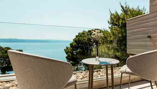 To stoler står ved et bord med en vase med blomster, med utsikt over havet og frodige trær. (AI-generert tekst)