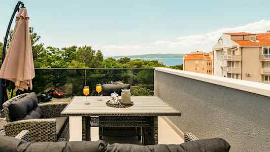 Terrasse med bord og stoler, oransje drikker i glass, parasoll og utsikt over trær, bygninger og hav. (AI-generert tekst)