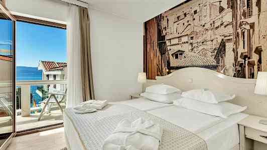 Et hotellrom med en dobbeltseng, et bord og en glassdør som fører til en balkong med utsikt over havet. (AI-generert tekst)