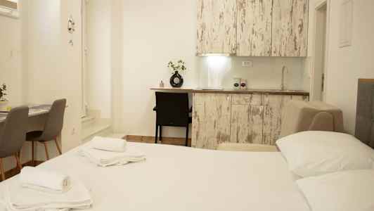 Bilde av et lyst innredet rom med seng, kjøkkenbenk og bord med stoler. (AI-generert tekst)