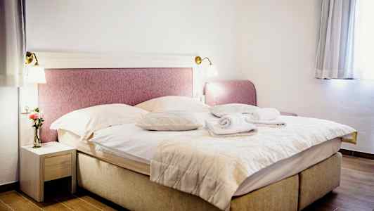 Et rom med en dobbeltseng oppredd med tepper og puter, to sammenrullede håndklær oppå. Et nattbord med en vase og en blomst til venstre og et vindu med gardiner til høyre. (AI-generert tekst)