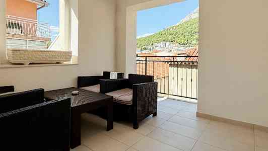 En terrasse med utsikt over tak og natur, møblert med mørke rottingstoler og bord. (AI-generert tekst)