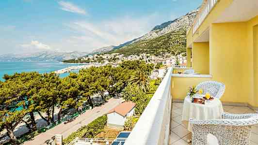 Utsikt fra en balkong på en gul bygning over en by ved havet og omkringliggende fjell. (AI-generert tekst)