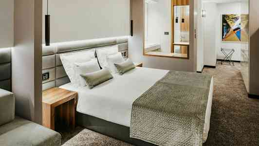 Et moderne hotellrom med en stor seng, hvite sengeklær, grått sengeteppe og puter, og en grå vegg-til-vegg teppe. (AI-generert tekst)