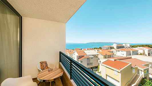 Utsikt fra en balkong med to stoler, et bord og utsikt over byområdet og havet. (AI-generert tekst)