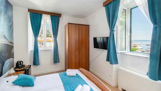 Hotellrom med dobbeltseng, blå gardiner og utsikt over en havn utenfor vinduet. (AI-generert tekst)