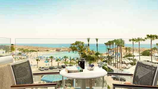 Utsikt fra en balkong med bord og stoler mot en strand med parasoller og palmer ved havet. (AI-generert tekst)