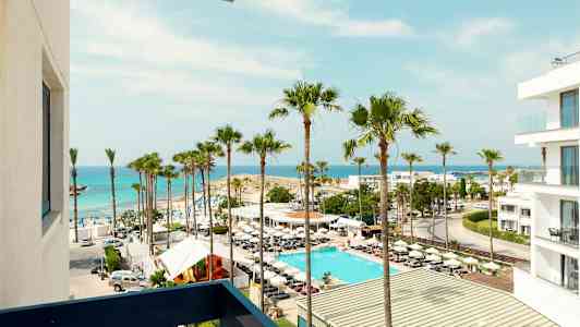 Utsikt over en strand med palmer, et basseng og en hvit bygning ved havet. (AI-generert tekst)