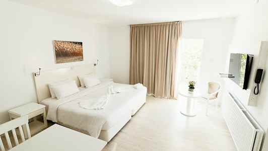 Et lyst og moderne hotellrom med en stor seng, hvite møbler og et bilde på veggen. Et bord med en blomst og en stol er synlig ved et vindu med beige gardiner. (AI-generert tekst)