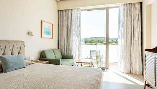 Et hotellrom med en seng, sofa og bord, samt en balkong med stoler og utsikt over grøntområder. (AI-generert tekst)