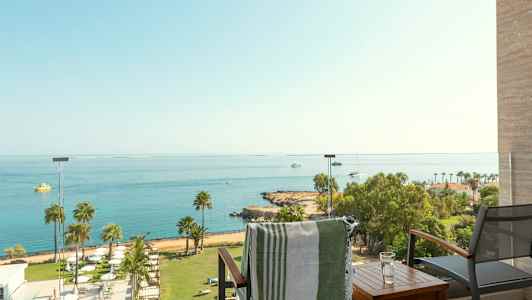 Utsikt fra en balkong mot havet med båter og en strand med palmer. (AI-generert tekst)