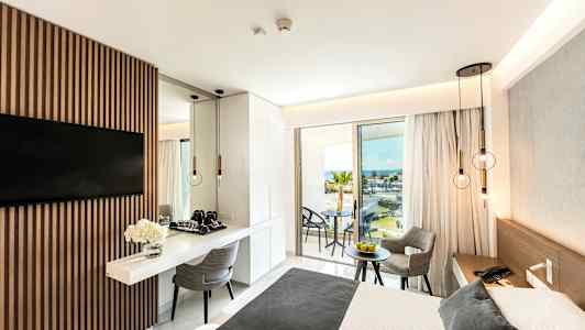 Moderne hotellrom med en dobbeltseng, bord og stoler samt utsikt over havet fra en balkong. (AI-generert tekst)