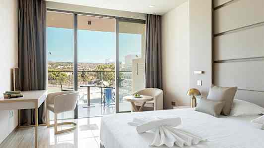 Et moderne hotellrom med en stor seng, et skrivebord og stoler, samt en balkong med utsikt. (AI-generert tekst)