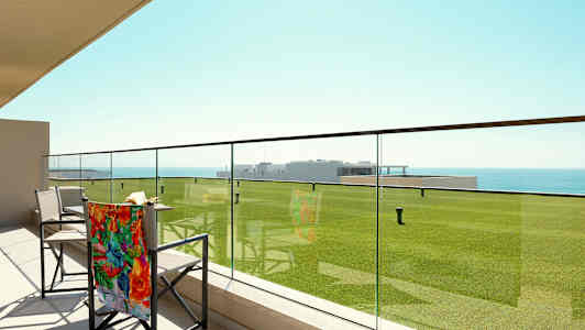 En terrasse med glassrekkverk, stoler og utsikt over grønne plener og havet. (AI-generert tekst)