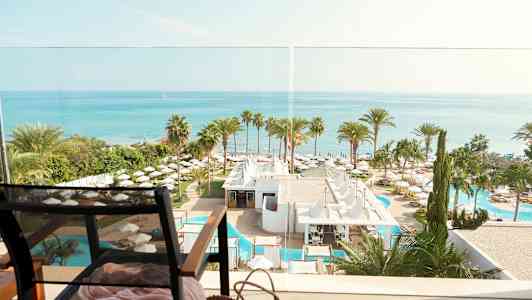 Utsikt over havet, hotellområde med palmetrær og bassenger, og en stol i forgrunnen. (AI-generert tekst)