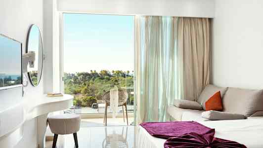 Et lyst hotellrom med en sofa, en seng med lilla teppe og en balkong med utsikt over grøntområde. (AI-generert tekst)