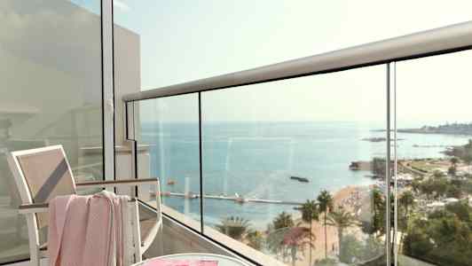 Utsikt fra en balkong mot havet, med en stol, et bord og et magasin. (AI-generert tekst)