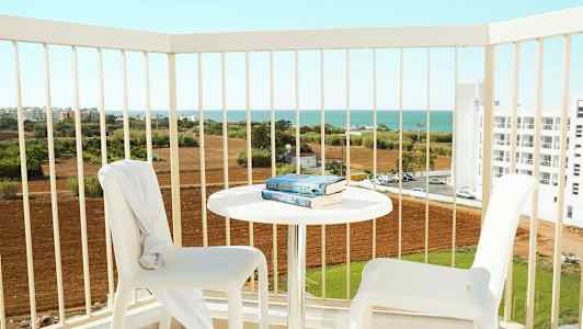 To hvite stoler og et bord med bøker på en balkong med utsikt over jordbrukslandskap og havet. (AI-generert tekst)