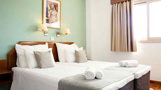 Interiør av et hotellrom med dobbeltseng, to sengelamper, et vindu med gardin og et dekorativt veggbilde. (AI-generert tekst)